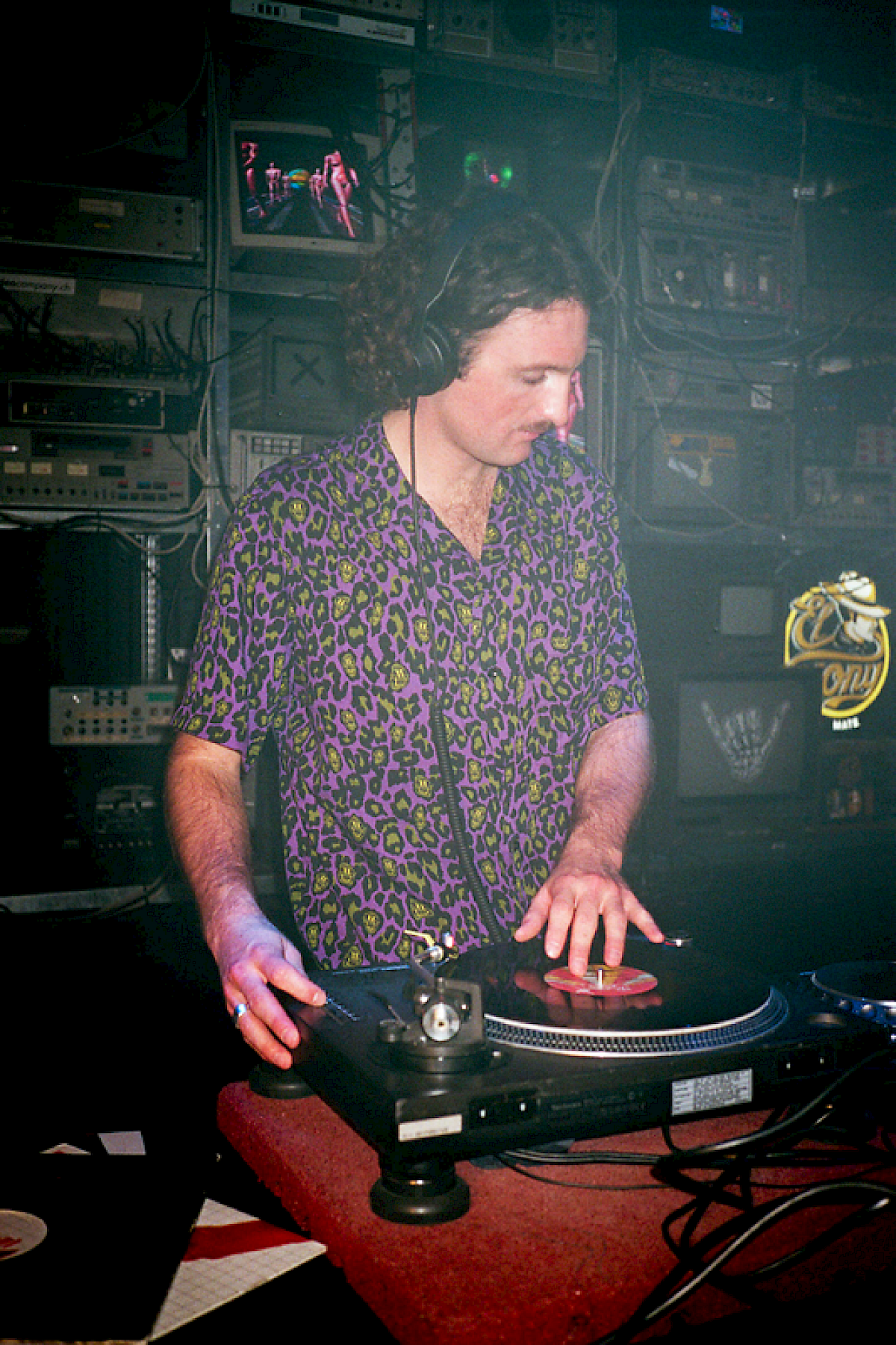 DJ, der Platten auflegt, trägt ein lila Leopardenmuster-Hemd in einer lebhaften Club-Umgebung.