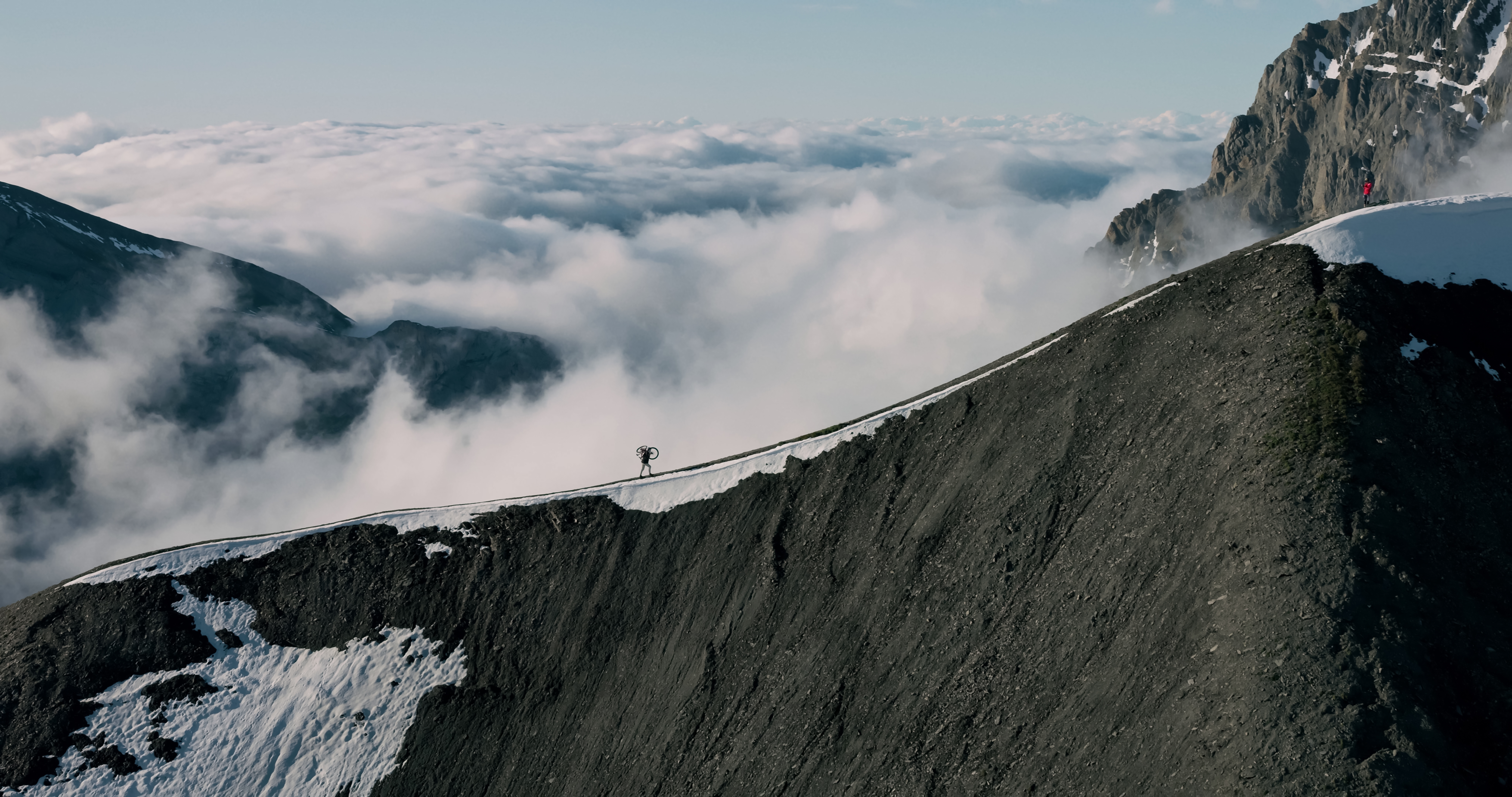 Eine Person trägt ein Fahrrad auf einem schneebedeckten Berggipfel über Wolken.