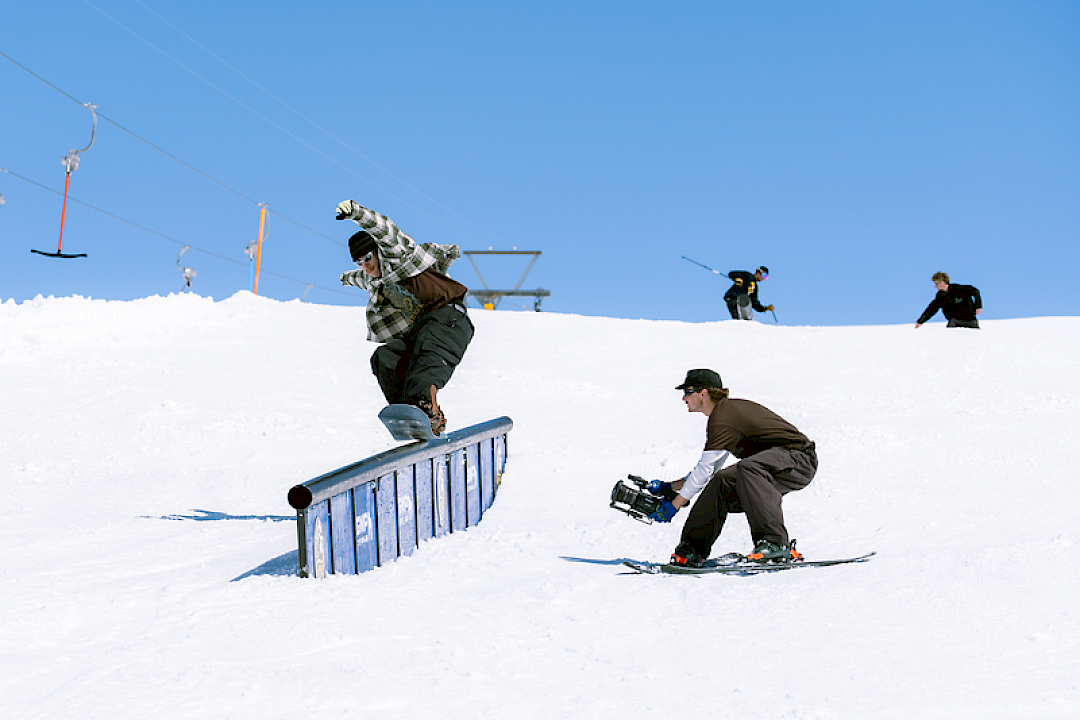 Snowboarder jumps on a rail while a cameraman films. Clear sky and snowy slope.