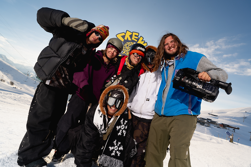 Group of young people in winter outfits, posing with a camcorder in a snowy mountain setting.