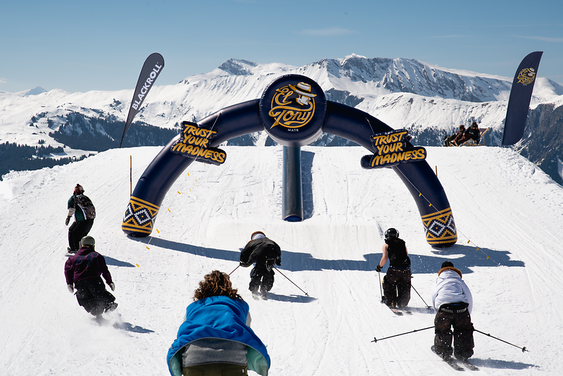 Skiers and snowboarders jump through an El Tony Mate arch on a snowy slope in the mountains.