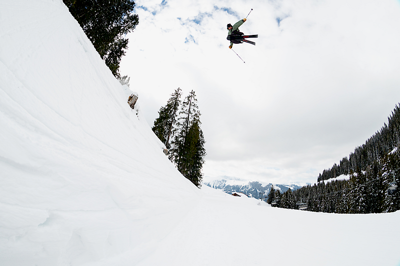 Skier jumps over a snowy slope in the mountains, surrounded by pine trees.