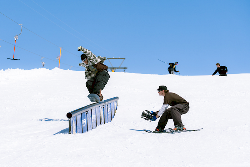 Snowboarder performs a trick on a rail while a cameraman records.