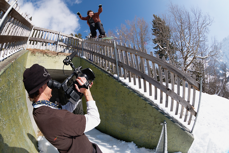 A snowboarder performs a trick on a railing while being filmed by a cameraman.