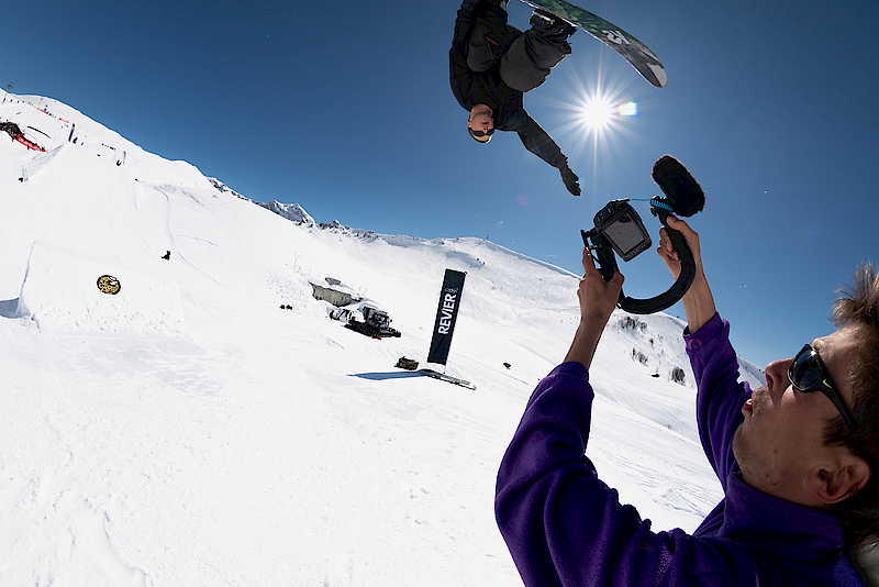 Snowboarder jumps in the air as a filmmaker captures him with a camera, mountains in the background.