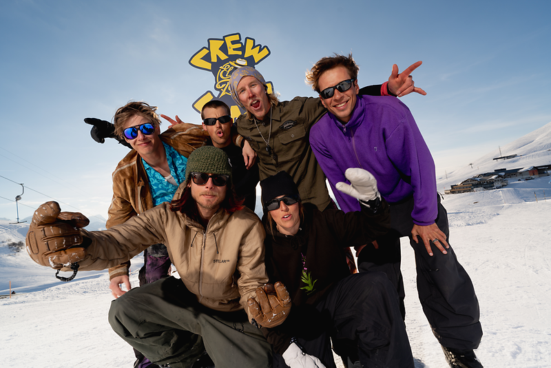 Six adventurous snowboarders happily pose in the snow with a large Crew sign in the background.