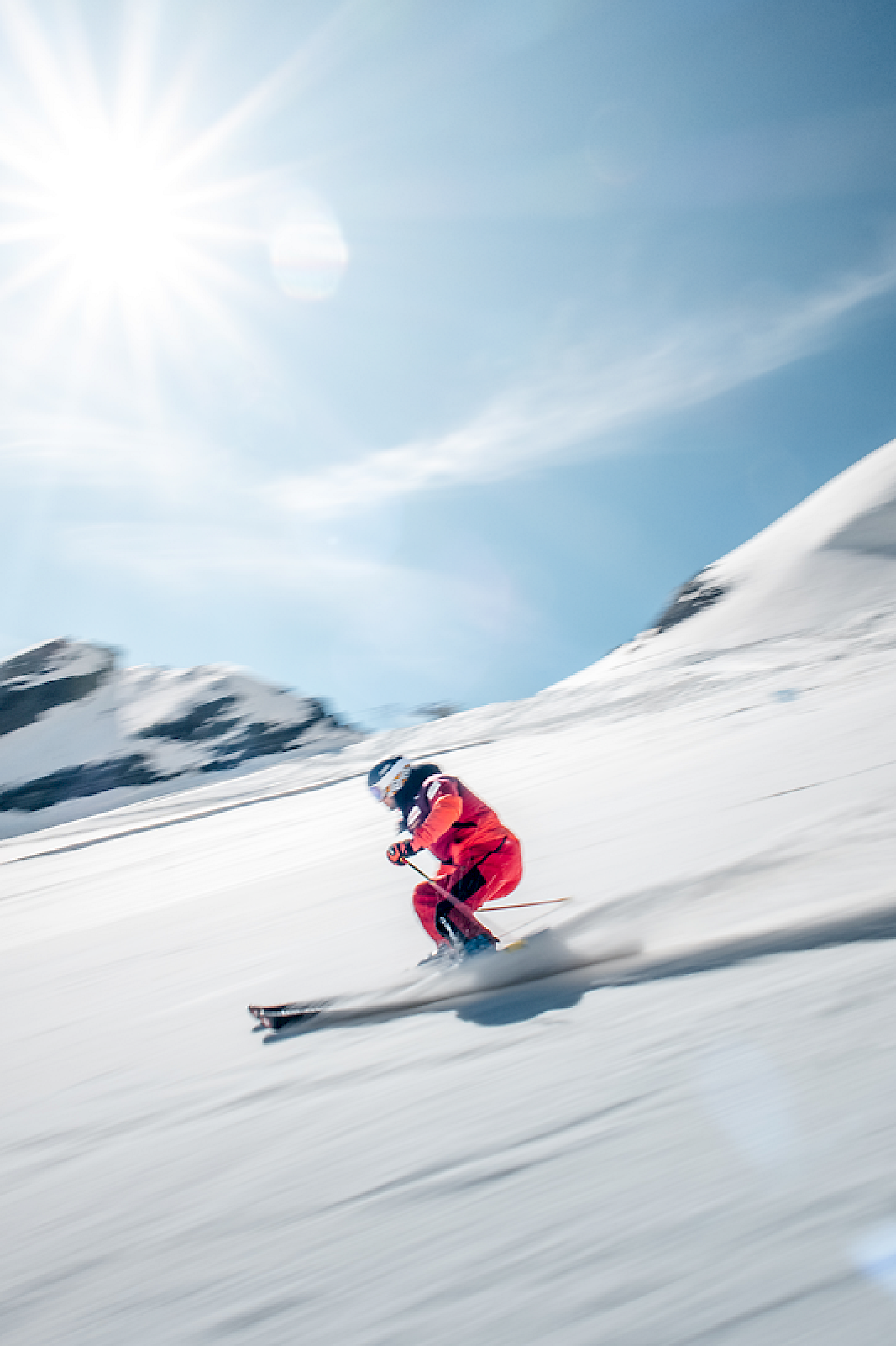 A skier in red gear speeds down the snow, with bright sun shining above.
