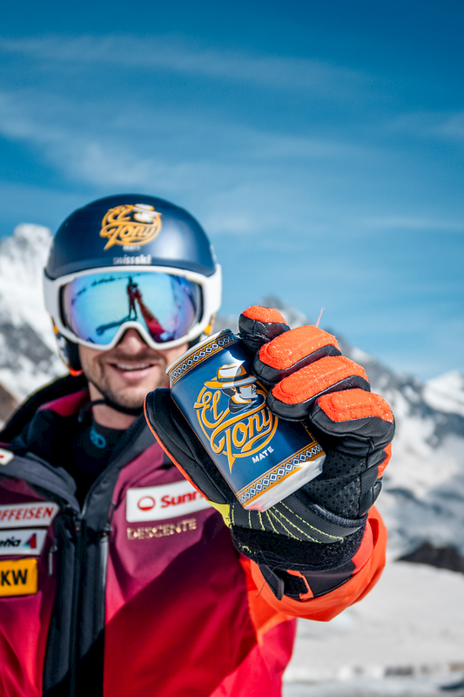 A skier holding an El Tony Mate can in the mountains, with a blue sky and snowy peaks in the background.