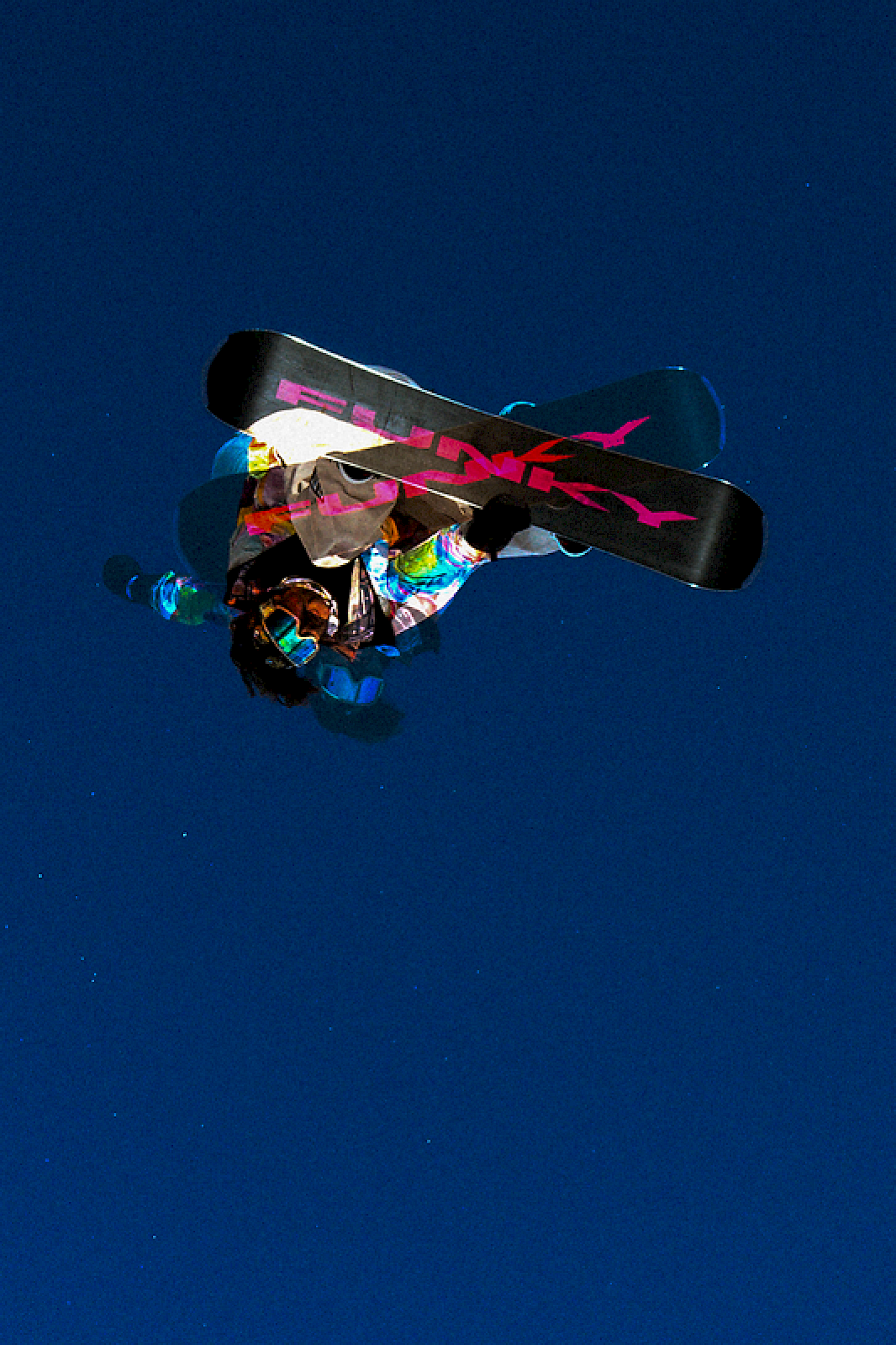 Snowboarder in de lucht met een kleurrijke outfit en een Funky snowboard tegen een blauwe lucht.