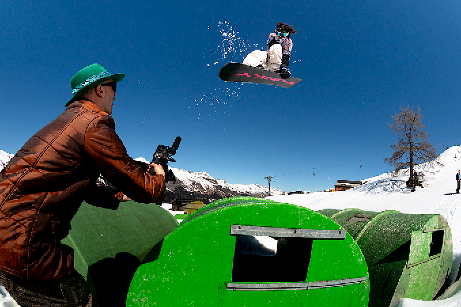 Een snowboarder springt over een groene ramp terwijl een toeschouwer met een camera opneemt.