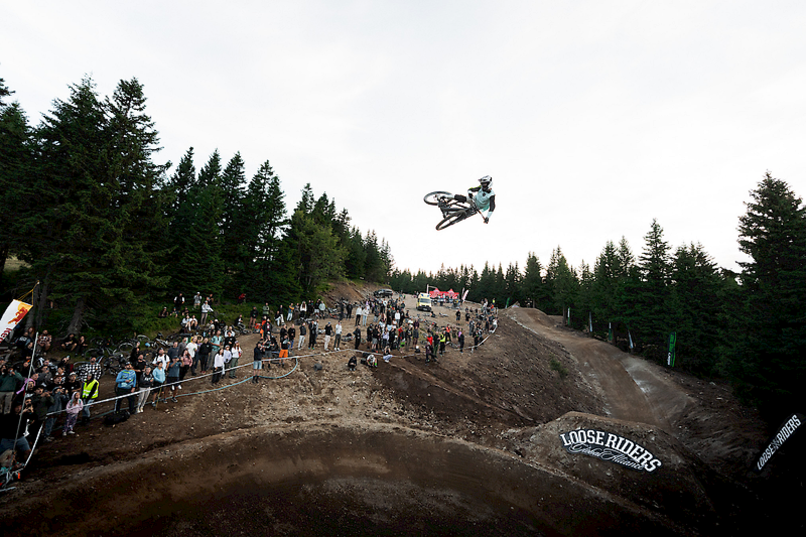Un rider di BMX salta sopra un trampolino davanti a una folla in un'area boschiva.