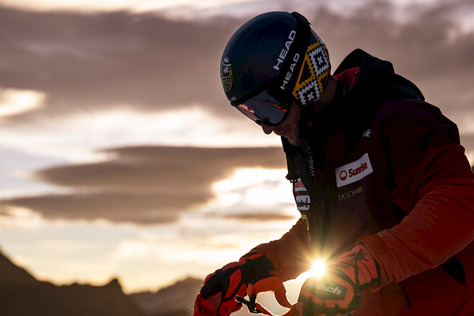 Skier bij schemering, zijn helm ajusterend met zonlicht op de achtergrond.