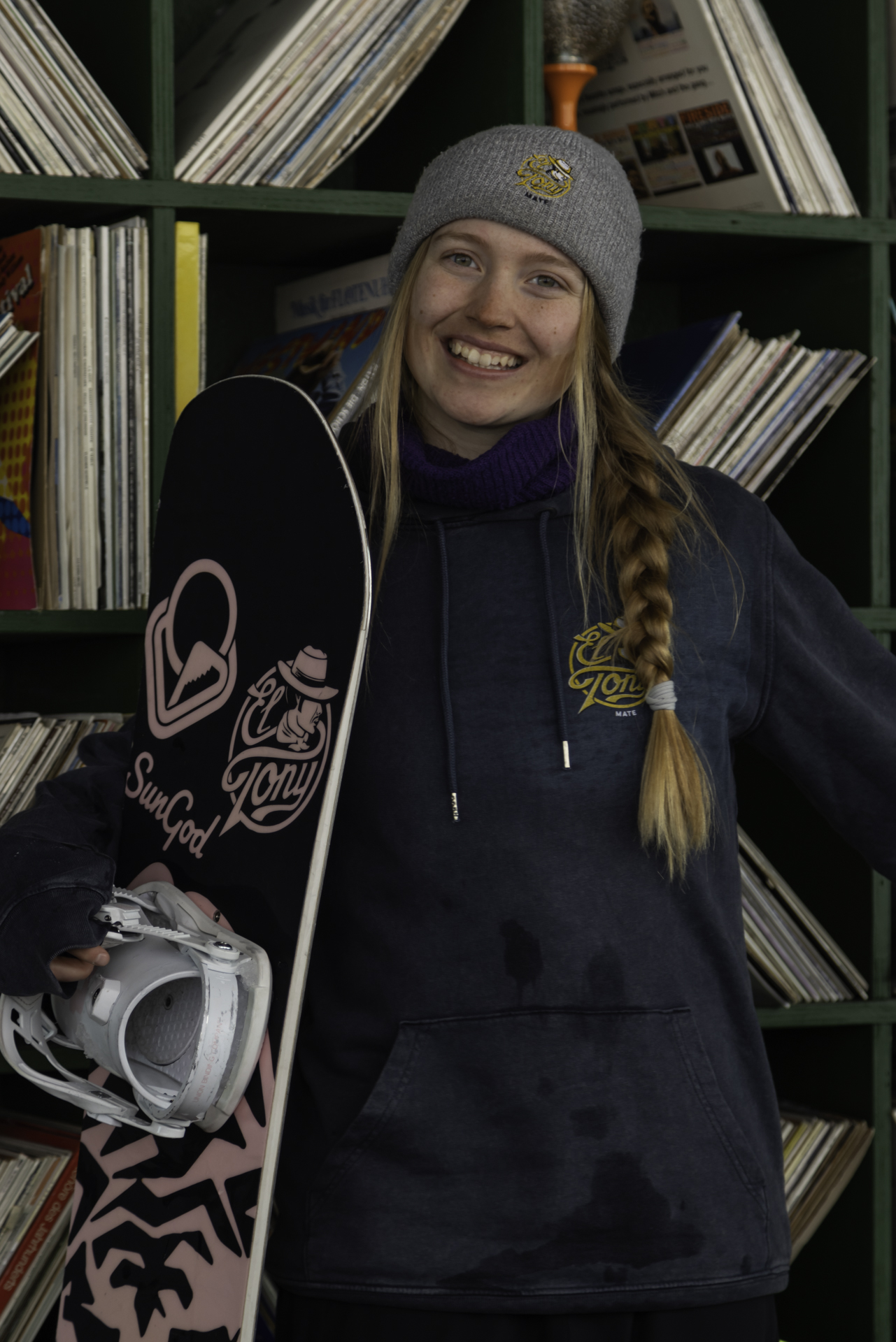 Smiling snowboarder with an El Tony Mate drink and snowboard in front of a vinyl record wall.