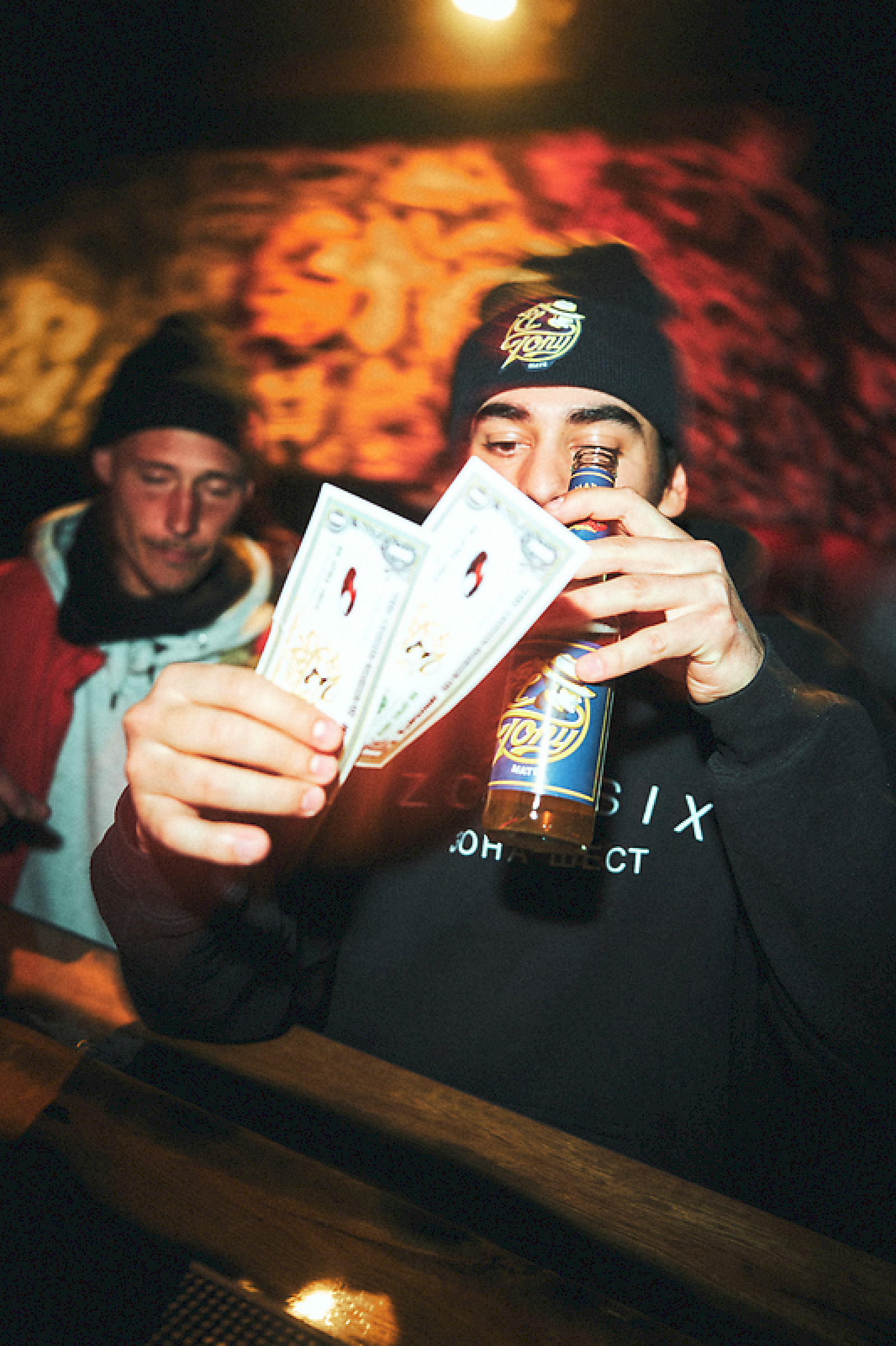 Junger Mann hält El Tony Mate-Flasche und Geldscheine in einer Bar, während jemand im Hintergrund zuschaut.