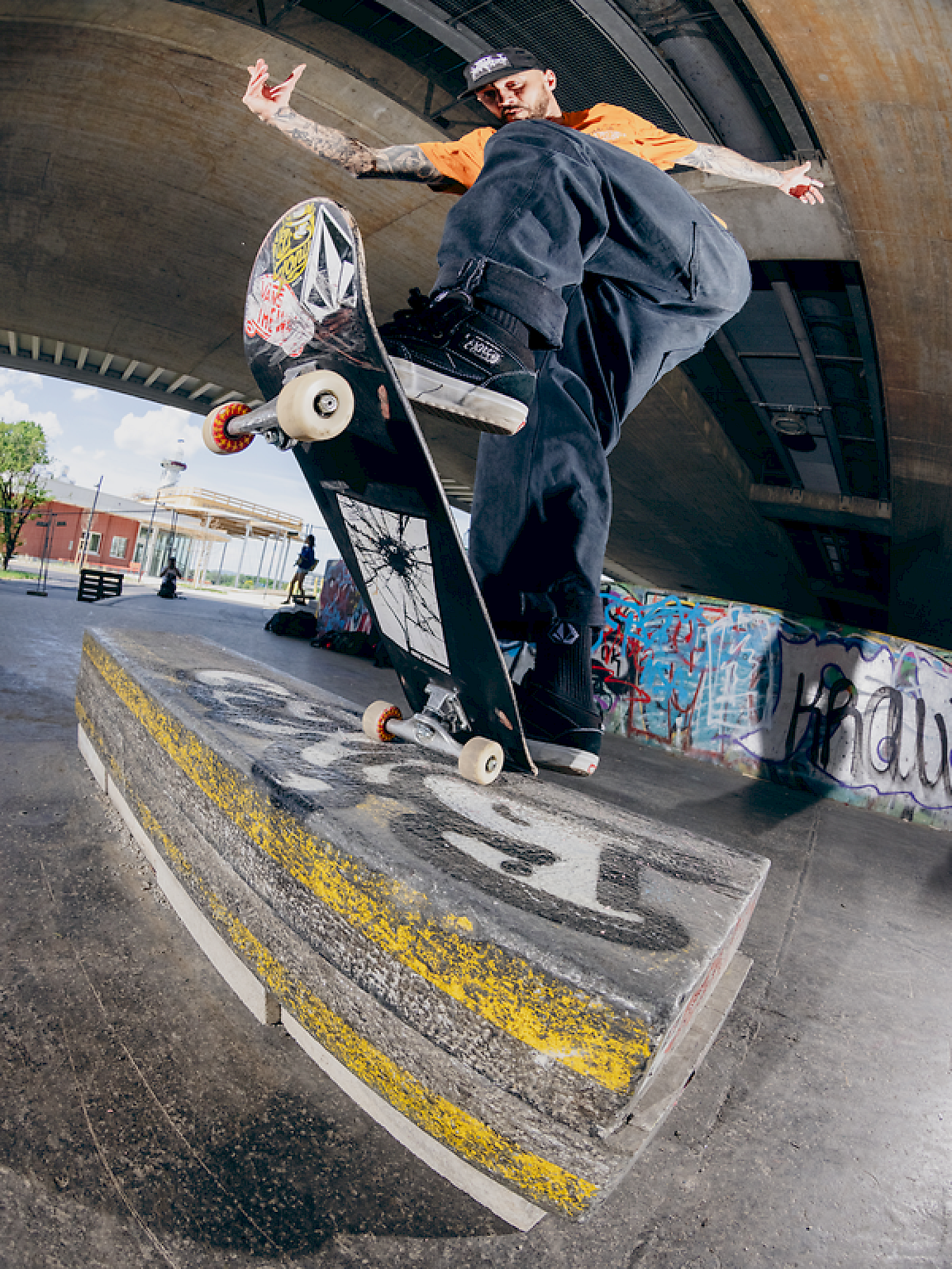 Skateboarder in orange shirt performs a trick over a graffiti-covered ledge.