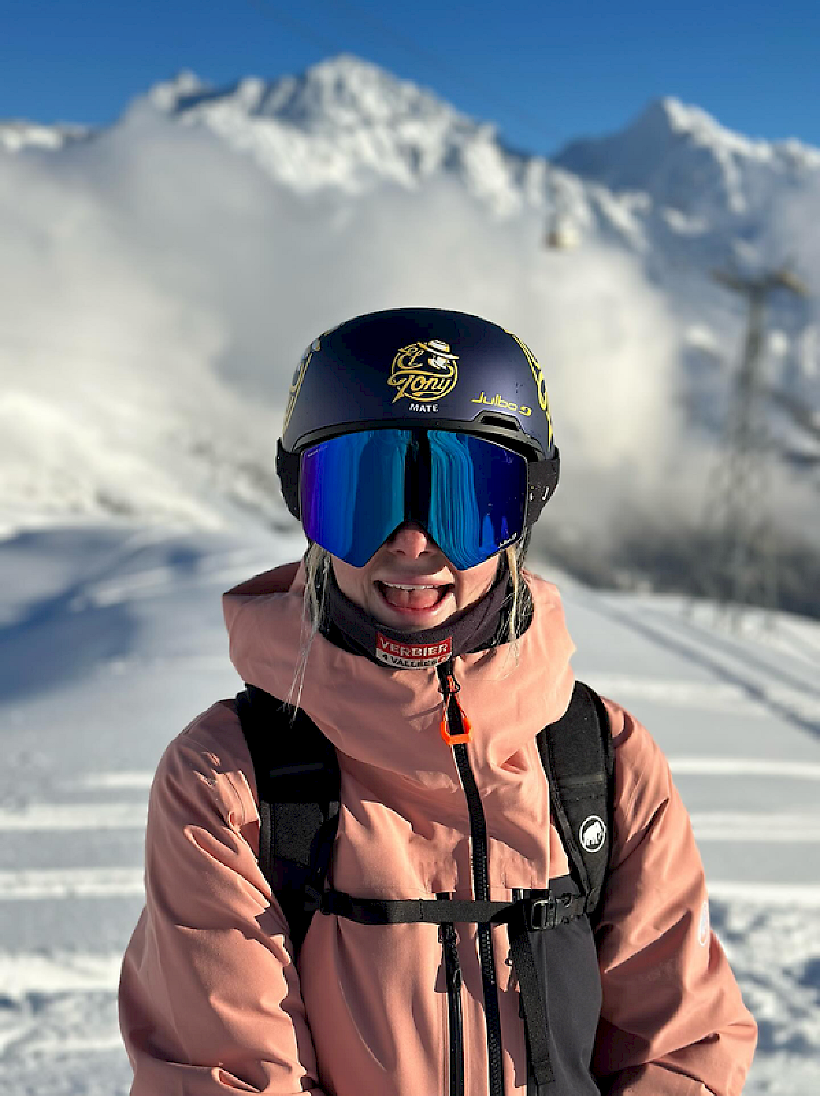 Frau mit Helm und Sonnenbrille im Schnee, lächelt, El Tony Mate Logo sichtbar.