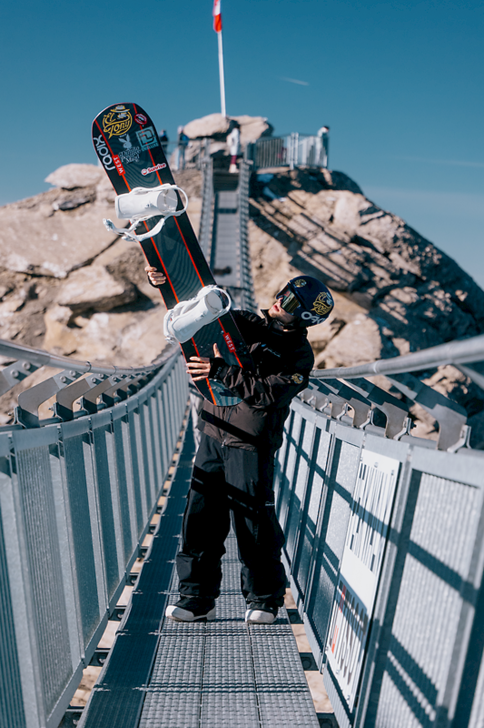 Snowboardfahrer hält sein Board auf einer Brücke mit Felsen im Hintergrund. Auf dem Peakwalk auf dem Glacier 3000