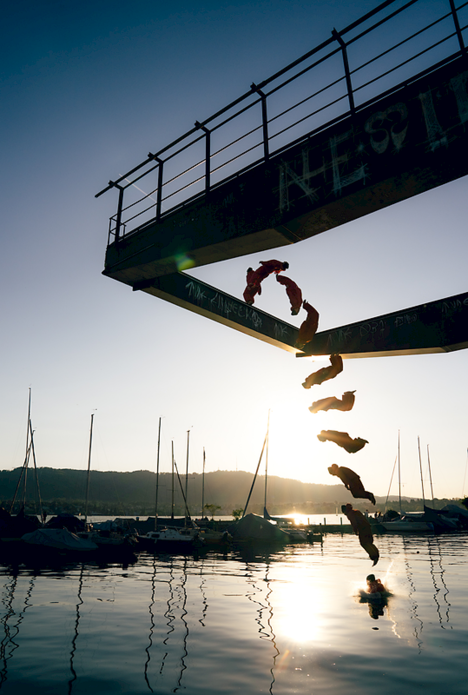 Person springt von einem Turm ins Wasser, umgeben von Booten und Sonnenuntergang.