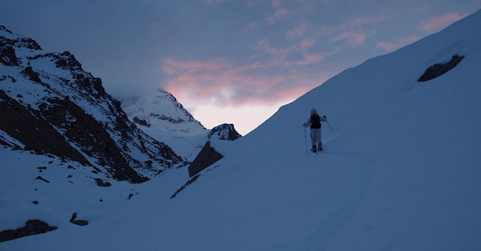 Ein Bergsteiger im Schnee mit El Tony Mate in der Hand, unter einem farbenfrohen Himmel.