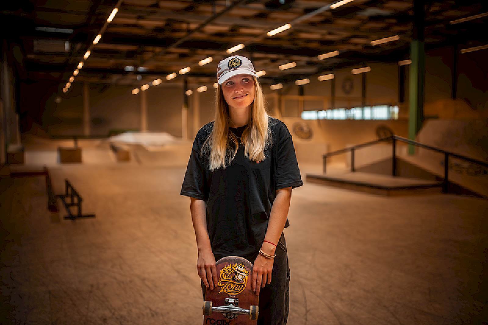 A skateboarder with an El Tony Mate cap stands in an empty skatepark.