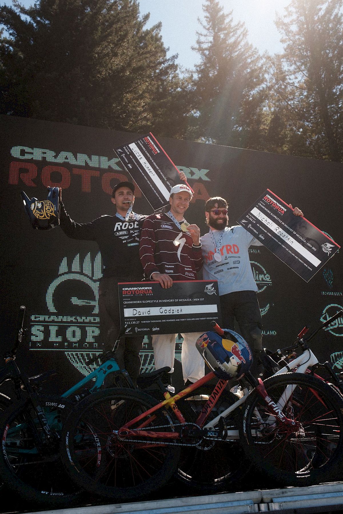 Drei Fahrer auf dem Podium mit Medaillen, Trophäen und Mountainbikes, ein fröhlicher Moment beim Rennen.
