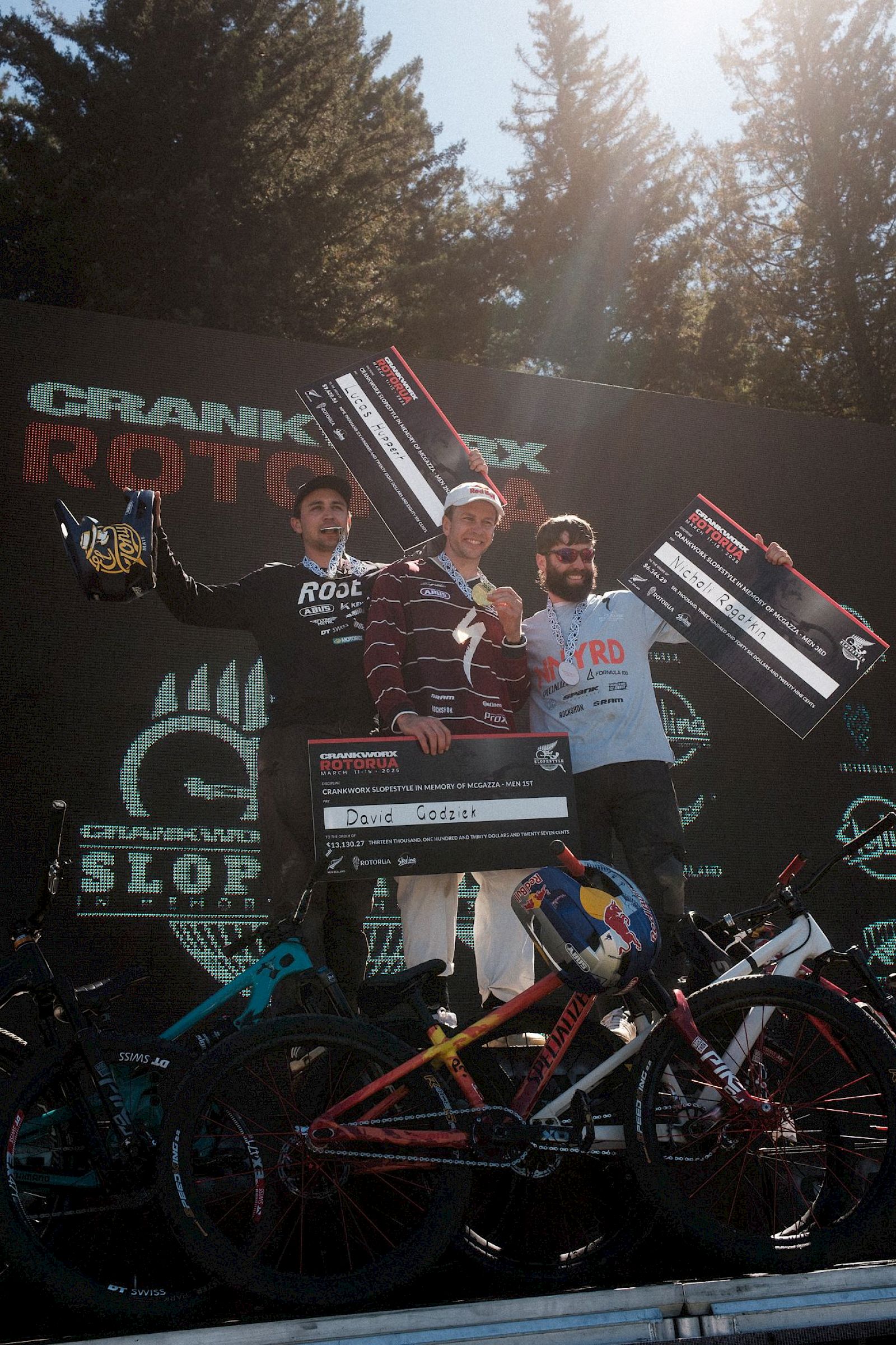 Drei Fahrer auf dem Podium mit Medaillen, Trophäen und Mountainbikes, ein fröhlicher Moment beim Rennen.