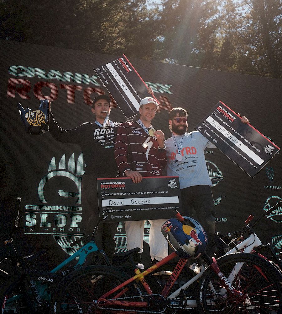 Three riders on the podium with trophies, enjoying a celebratory moment after the race, bikes in front.