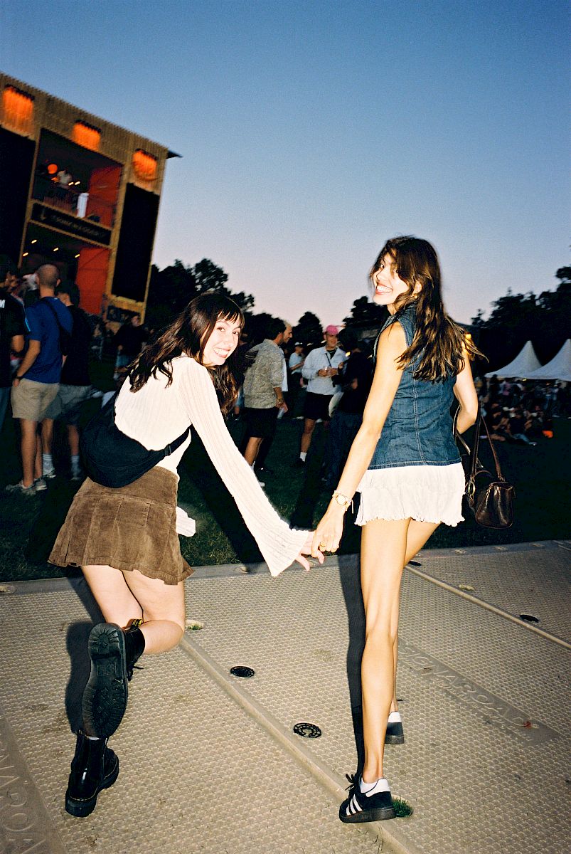 Two friends hold hands at a festival, people and stalls in the background as dusk falls.