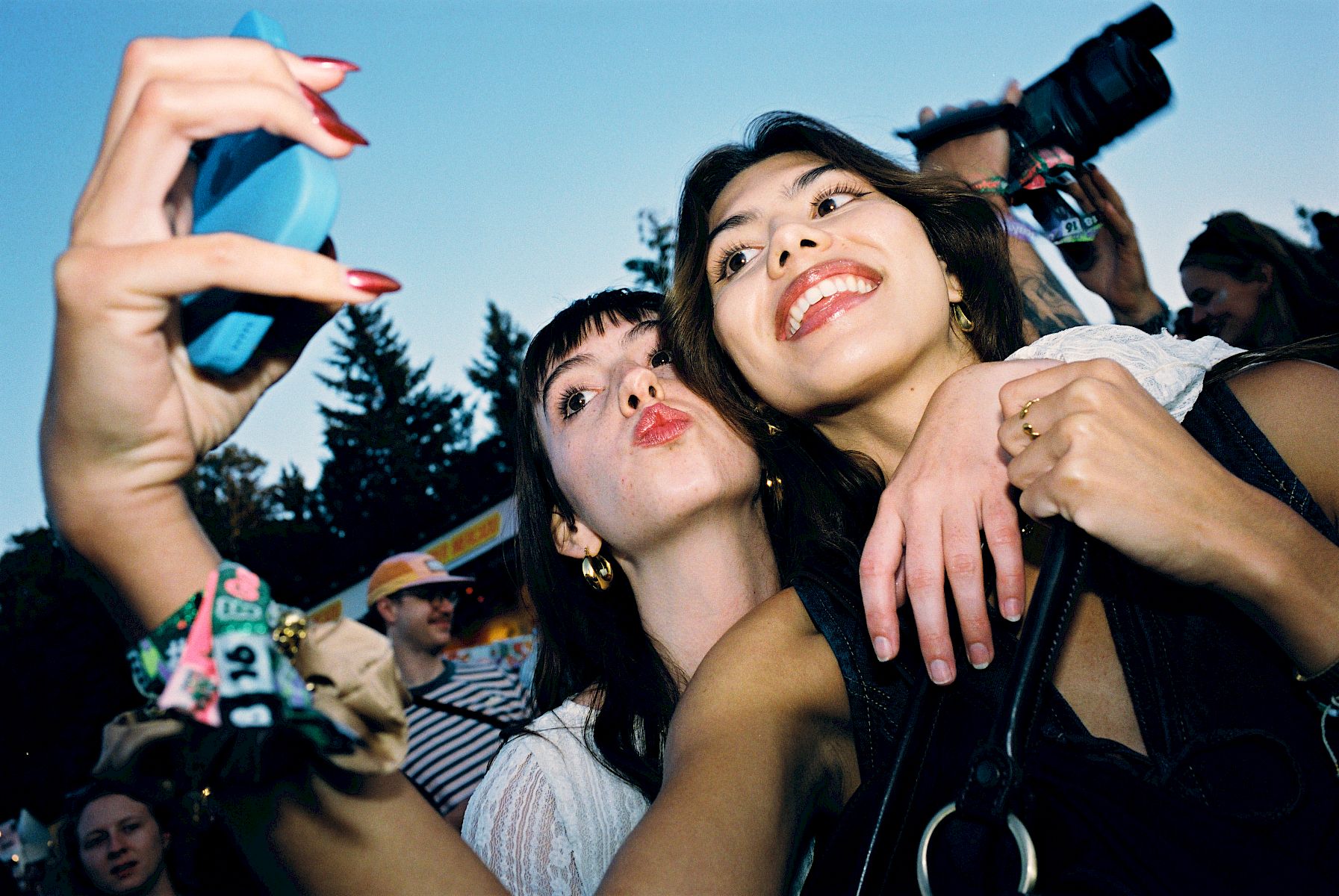 Two women take a selfie at a festival, with people and an El Tony Mate bottle visible in the background.
