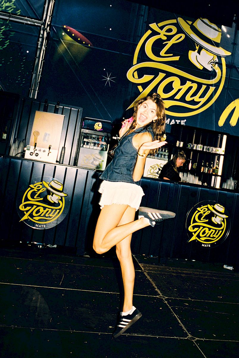 Young woman joyfully poses in front of El Tony Mate booth at a festival.