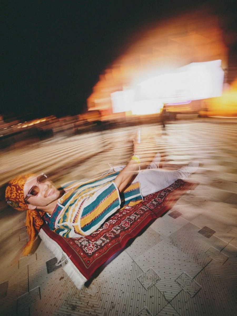 Man in colorful shawl relaxes on a carpet, enjoying El Tony Mate in a lively setting.