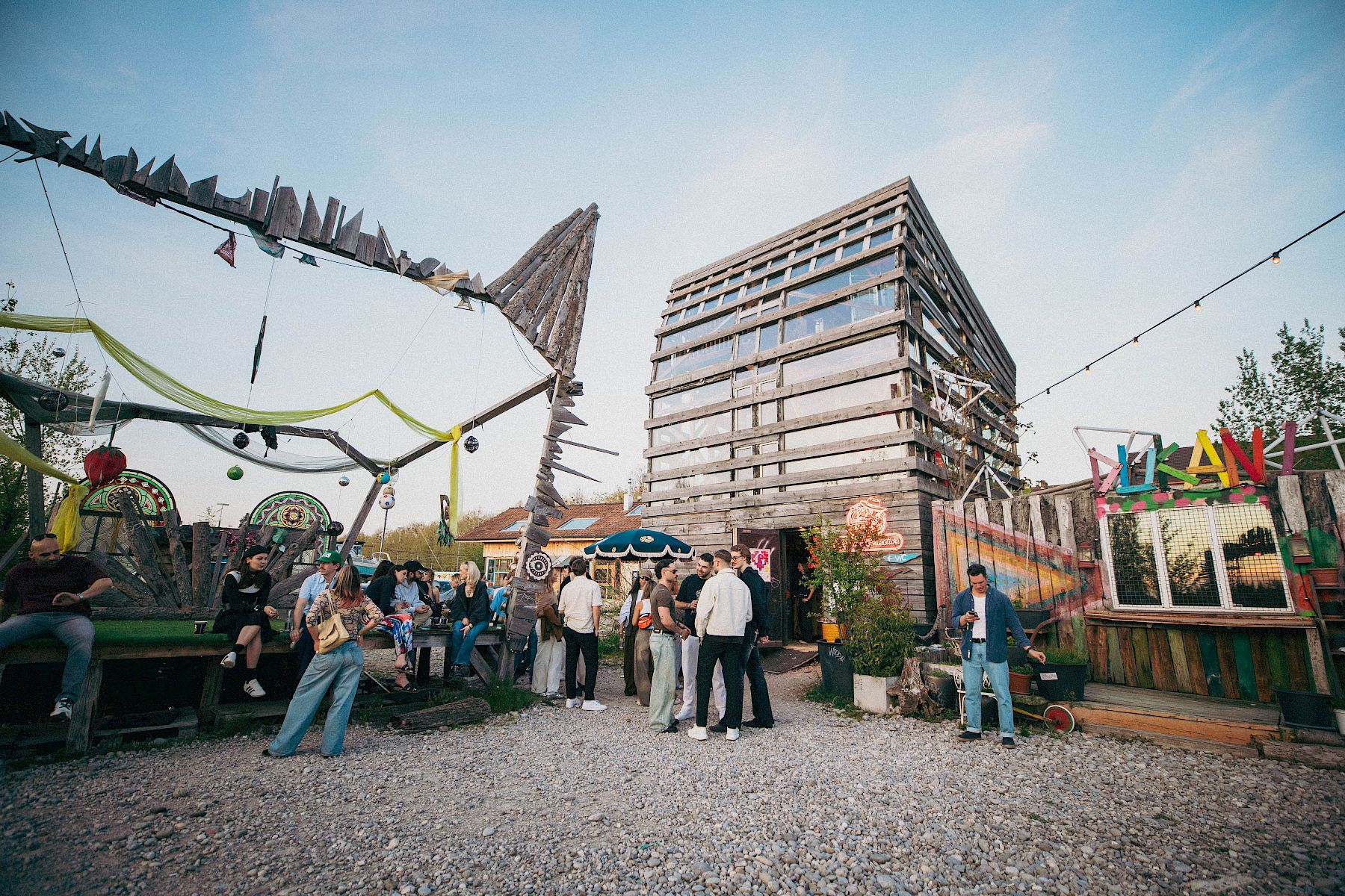 Luogo all'aperto vivace con persone, bancarelle colorate e un moderno edificio in legno sullo sfondo.