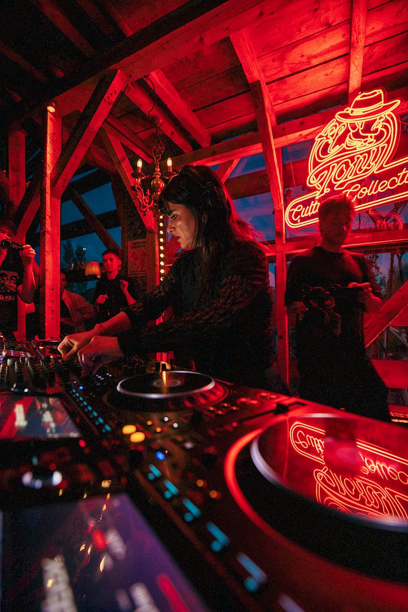 DJ che si esibisce sotto un'insegna al neon El Tony Mate, persone che ballano sullo sfondo.