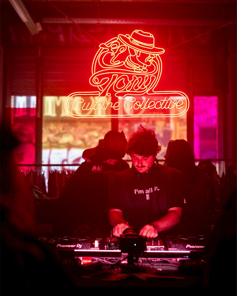 Een DJ die op El Tony Mate draait, omringd door neonlichten en een levendige culturele sfeer.