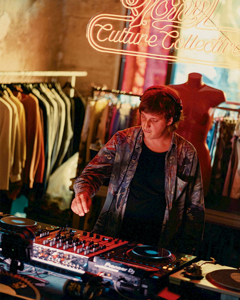 DJ aan het mixen in een levendige kledingwinkel, met neonlichten op de achtergrond.