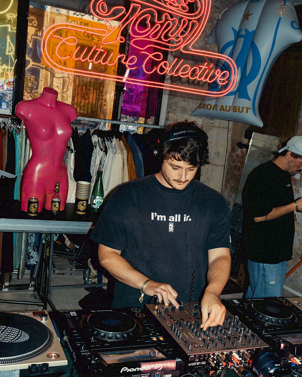 Een DJ mixt muziek bij zijn booth, omgeven door drankjes en een levendige, artistieke sfeer.