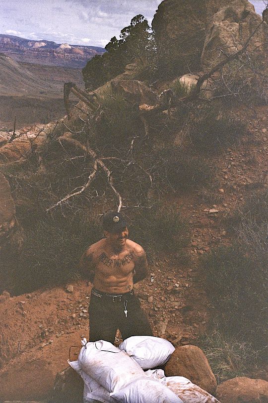 Un uomo tatuato sta su un terreno roccioso con sacchi e alberi sullo sfondo.