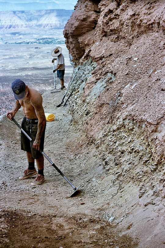 Due lavoratori scavano in un paesaggio roccioso, uno tiene un attrezzo.