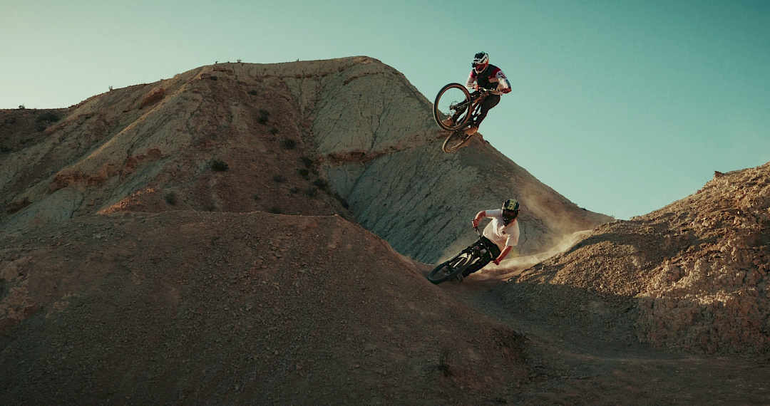 Twee mountainbikers springen over een heuvelachtig landschap met stoffige, droge aarde.