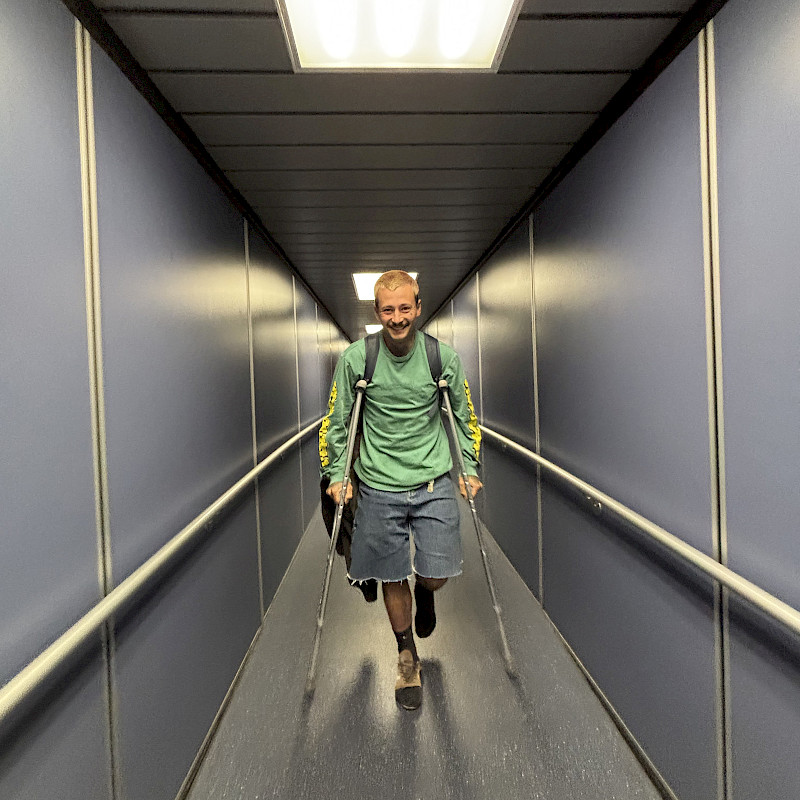 Un uomo con le stampelle cammina nel corridoio dell'aereo, indossando una maglietta verde e pantaloni corti.