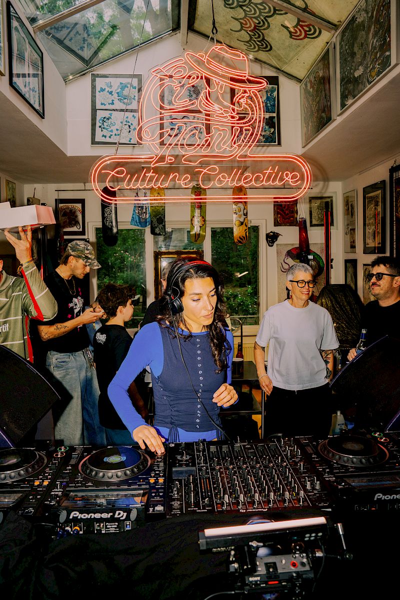 DJ en performance dans un espace animé avec enseigne néon El Tony Mate, entouré de gens et d'art.