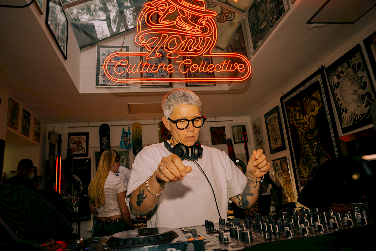 DJ at the mixer in a creative space featuring 'El Tony Mate' and colorful artwork in the background.