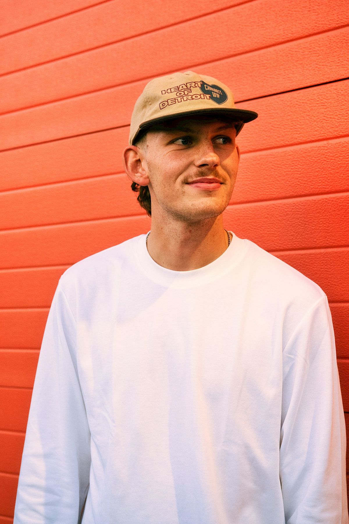 Un jeune homme en sweat-shirt blanc et casquette devant un mur rouge.
