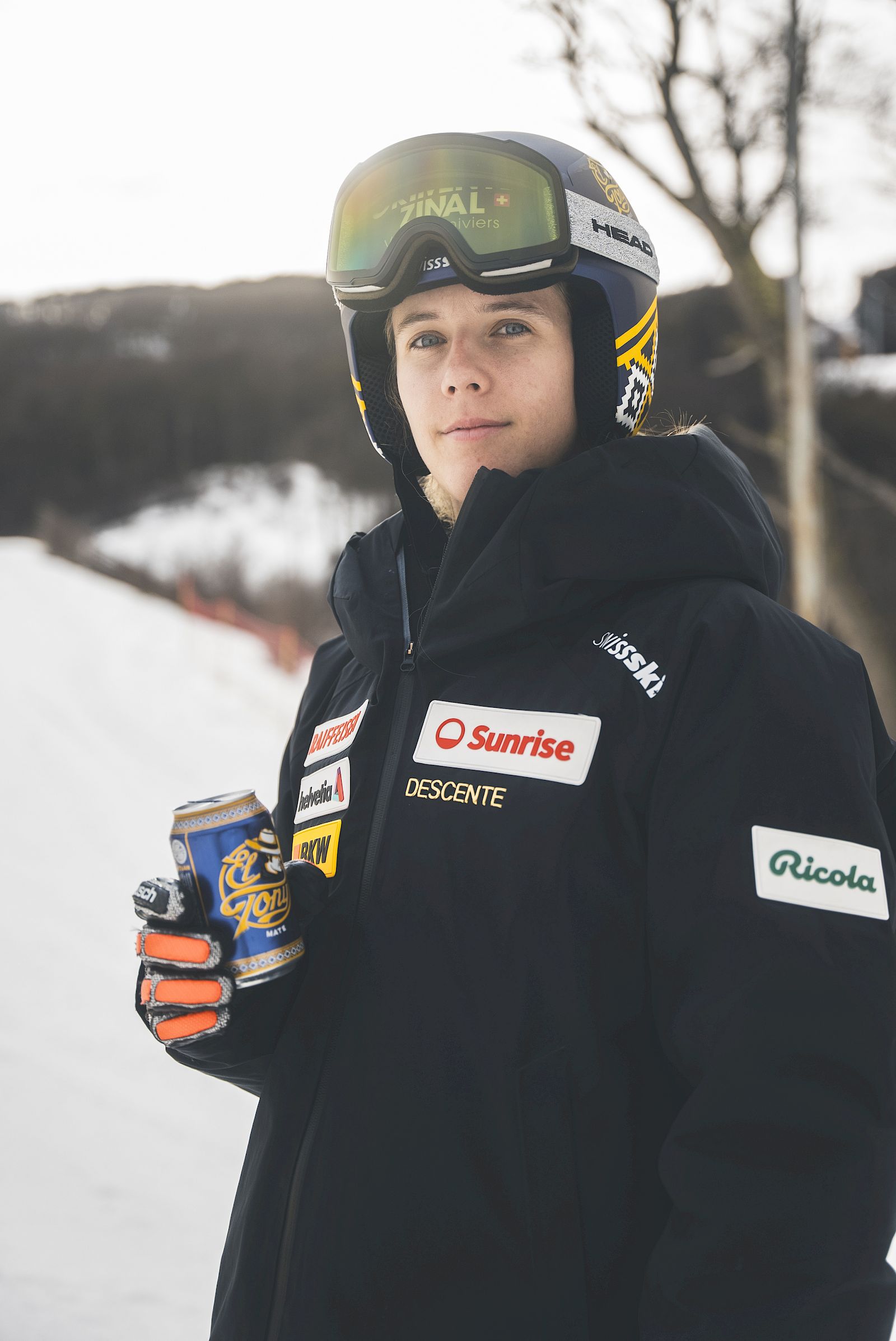 Junger Skifahrer in Schnee mit El Tony Mate Getränk in Hand, trägt Skikleidung und Sonnenbrille.