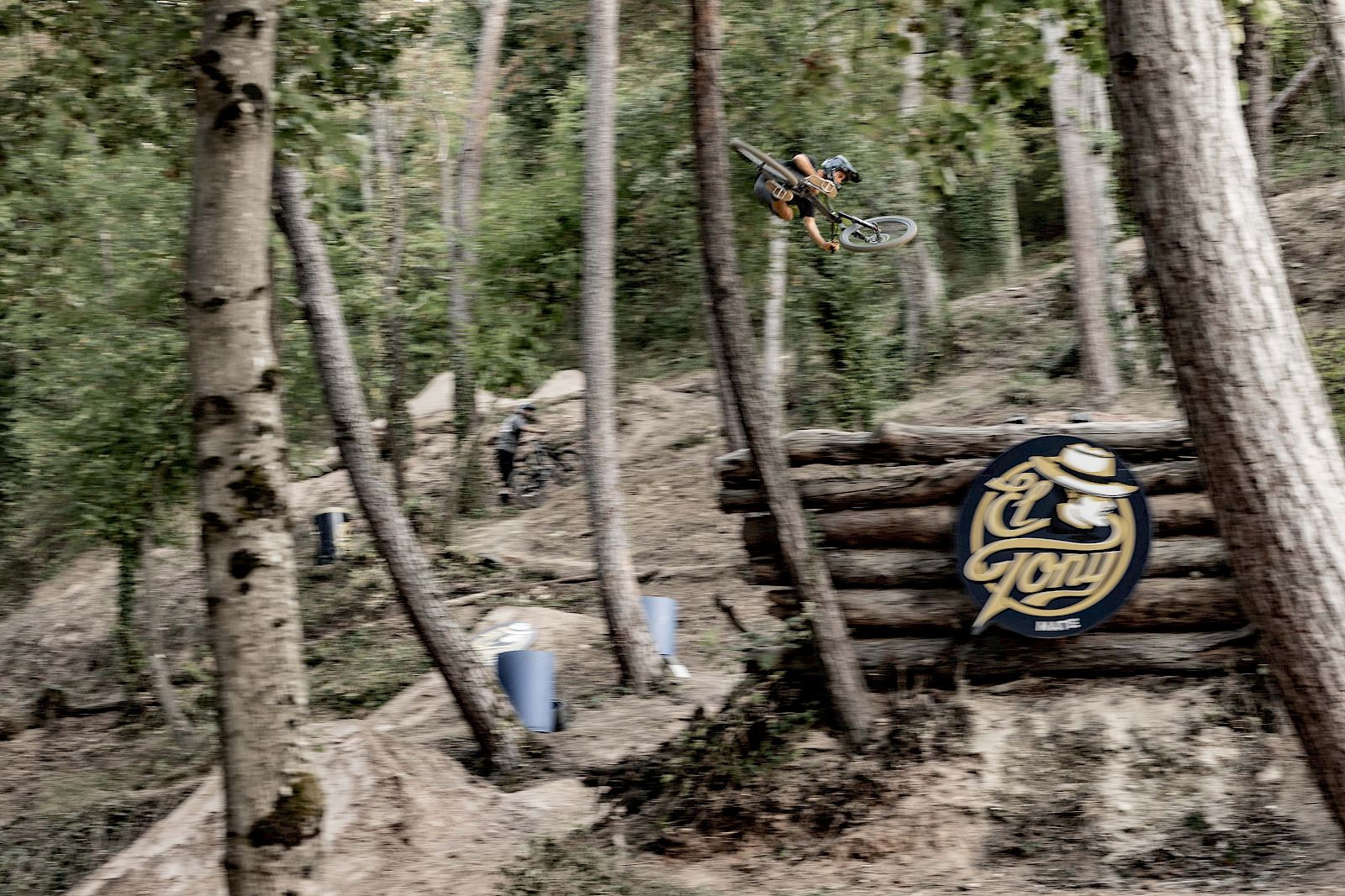 Un cycliste BMX saute par-dessus une structure en bois devant un panneau El Tony Mate dans une forêt.