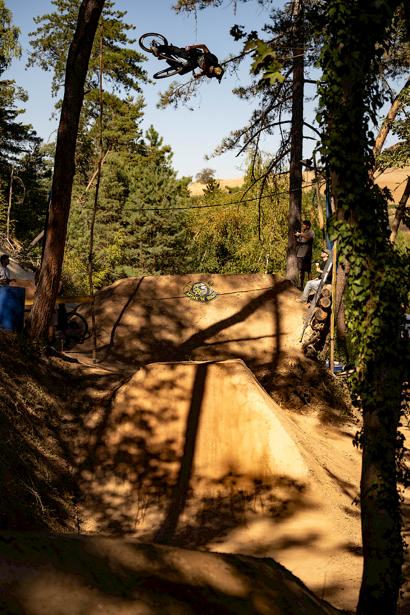 Un cycliste BMX saute par-dessus une rampe avec des arbres et des spectateurs en arrière-plan.