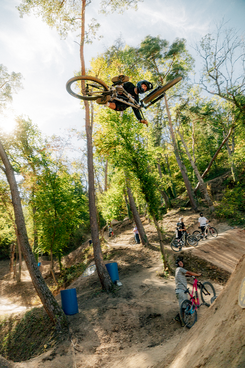 Un garçon fait un saut en BMX au-dessus d'une colline dans une forêt, d'autres riders l'observent.